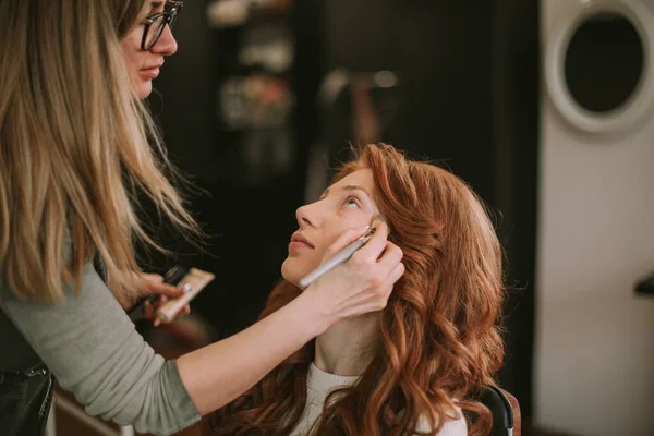 ブロンド作るアーティスト置くいくつかの顔の粉オン彼女の赤毛クライアント上のザ化粧サロンとともにAメイクアップブラシ — ストック写真