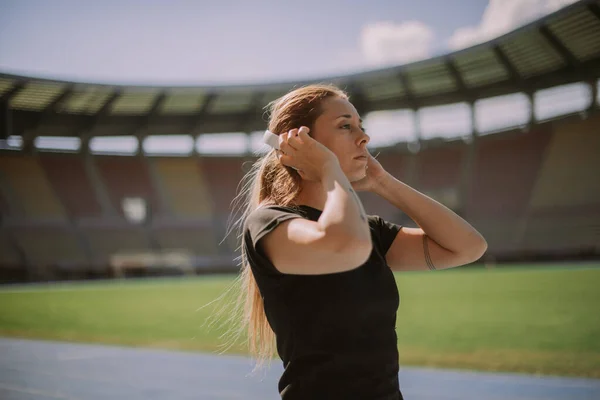 Beautiful female sportsperson is about to run on a stadium\'s track. She is putting her headphones on