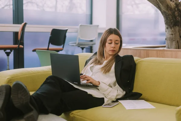 在同事工作的地方 坐在黄色沙发上打字和在笔记本电脑上工作的金发女商人的近照 — 图库照片
