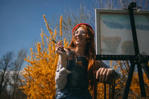 黄色の花の前の公園に座っている間 生姜の女の子の笑顔と彼女のキャンバスに絵を閉じます — ストック写真
