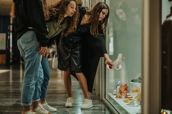 女孩们看着购物中心的商店橱窗 布鲁内特姑娘指着她想买的高跟鞋 — 图库照片