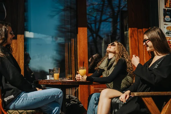 女性の友人が楽しい会話と笑顔をしている間 豪華な巻き毛の女の子は太陽の光線を楽しんでいます 彼らはコーヒーバーに座っている — ストック写真