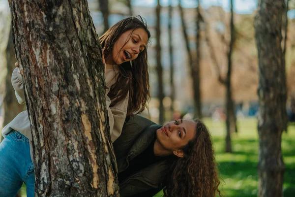 两个可爱的女孩躲藏在公园的树后 她的前景被拍摄到了 布鲁内特女孩看着她卷发的朋友 — 图库照片