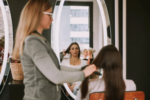在美发店梳头时 漂亮的黑发姑娘看着镜子 与理发师交谈 — 图库照片