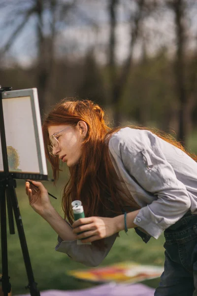 美丽的生姜姑娘站在公园边 在画架上画了一幅画 还创作了一幅五彩斑斓的杰作 — 图库照片