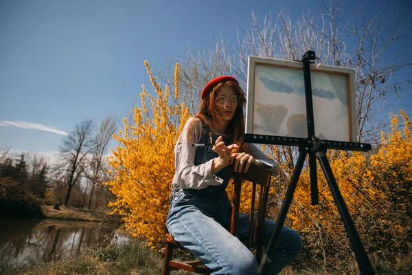 公園の黄色の花の前に座っている間 キャンバス上の絵を見て美しい生姜の女の子 — ストック写真