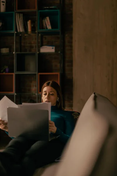 stock image Young girl lying on the sofa with the documents in her hand and reading from the laptop at the cafe bar