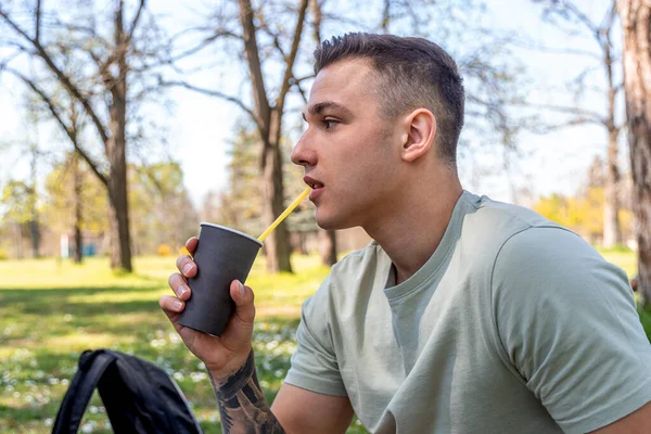 公園に座っているハンサムな男の側面図 紙コップからコーヒーを飲むと離れて見て — ストック写真