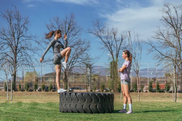 トラックのタイヤの上に立っている間グレーのスポーツウェアの練習足でブルネットの女の子 彼女の友人は彼女を見ている — ストック写真