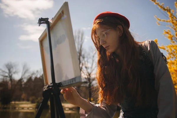湖の近くの公園のキャンバスに赤い髪の眼鏡と赤い帽子の絵を身に着けている美しい女の子 — ストック写真
