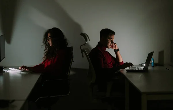 两名年轻同事深夜在一间黑暗的办公室工作的侧景照片 — 图库照片