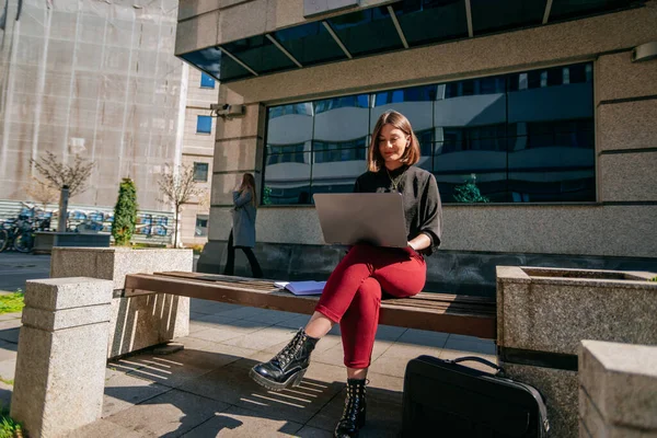 Two business girls working remotely outdoors. Brunette woman sitting on a bench and working on the lap top while her colleague is having a phone call in the back