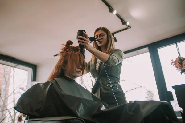 金发美发师在美发厅吹干她客户的红头发 — 图库照片