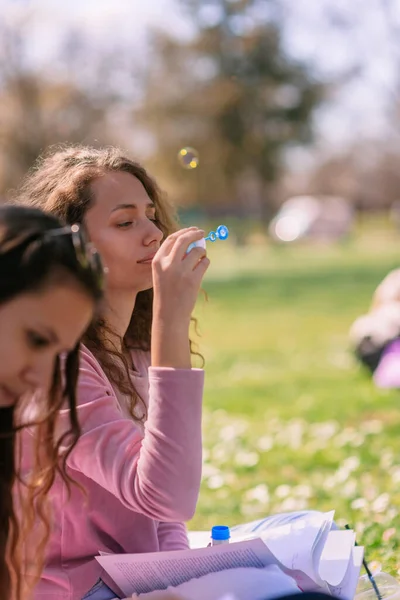 两个女学生在公园里当她的朋友在准备考试时 卷发女孩在吹泡泡 — 图库照片