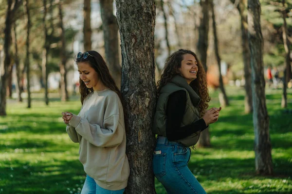 喜形于色的年轻姑娘们靠在公园的树上 布伦内特姑娘望着花朵 一边看着她的朋友一边笑 一边和她的朋友说话 — 图库照片