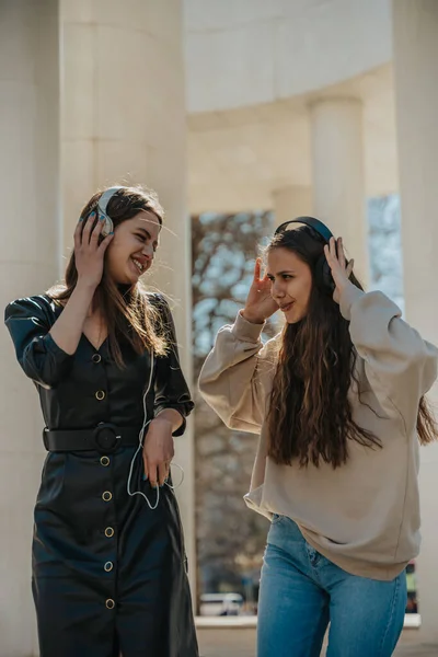 Stock image Beautiful brunette girl (left) listening to music on her headphones, singing and looking at her friend with slight smile on her face. Brunette girl in black overcoat smiling