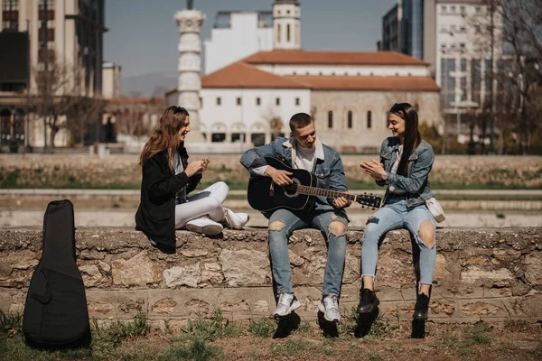 ハンサムな男の子はギターを弾いている間に彼の二人のガールフレンドと川の近くに座って — ストック写真