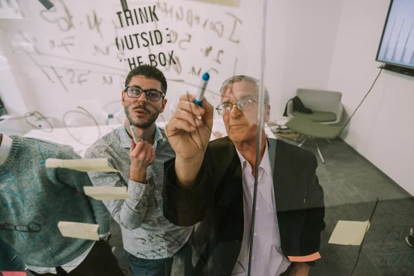 Close up of older business man pointing at the glass with his markers and talking with his younger colleague at co working space