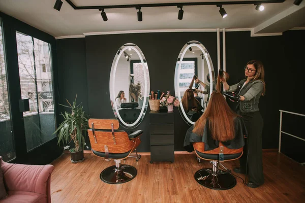 美发师在美发厅吹干她客户的红头发 而另一个客户在等她 — 图库照片
