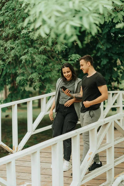 在公园的白桥上散步时 这对美丽的夫妇一边看着电话 一边看着对方 — 图库照片