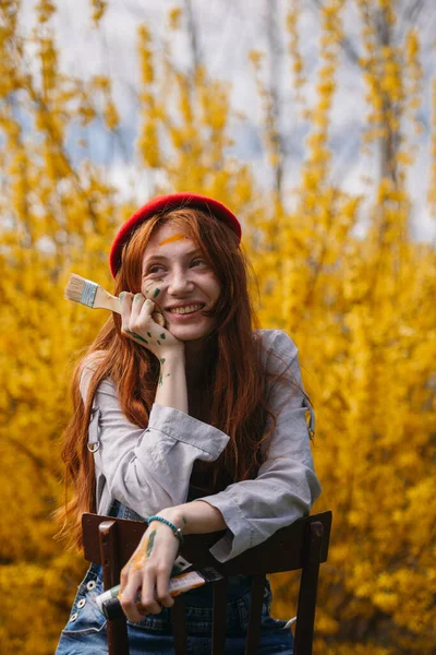 黄色の花の前に座って笑顔ながら離れて見て彼女の顔にペイントと美しい女性画家の肖像画 — ストック写真