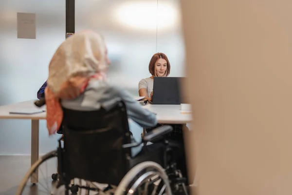 漂亮的女孩在办公室最上面的膝盖上工作 坐在轮椅上的残疾穆斯林女性坐在她的面前 — 图库照片