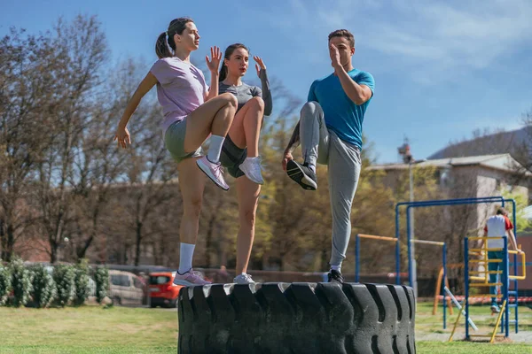 公園の巨大なトラックのタイヤの上に立っている間に彼らの足を行使する3つのスポーツの友人のショット — ストック写真