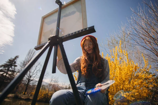 湖の近くの公園で笑顔に座っている間にキャンバスに美しい生姜の女の子の絵 — ストック写真
