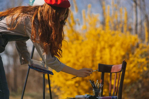 公園に立っている木製の椅子にガラスからブラシを取っている生姜の女の子の画家のクローズアップ — ストック写真