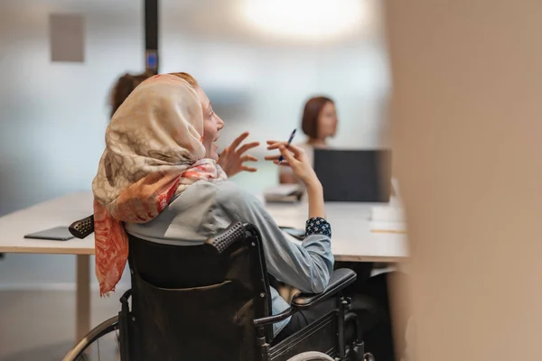 坐在轮椅上的年轻残疾穆斯林女工向她的同事解释一些事情 — 图库照片