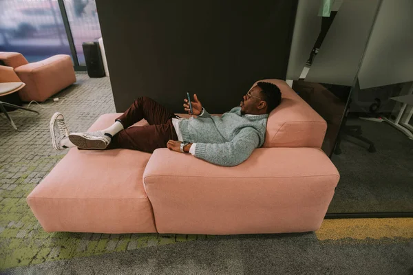 ピンクのソファの上に寝そべってるハンサムなビジネスマンとコワーキングスペースで電話を見て — ストック写真