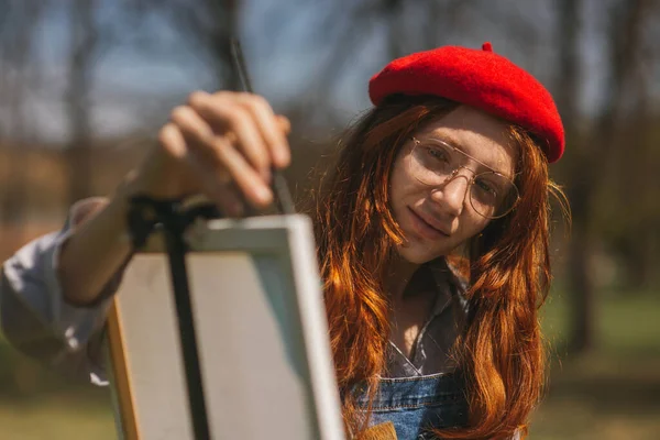 公園に立っている間 キャンバスの上に赤いベレー帽の絵を描いた美しい生姜の女の子のクローズアップ — ストック写真