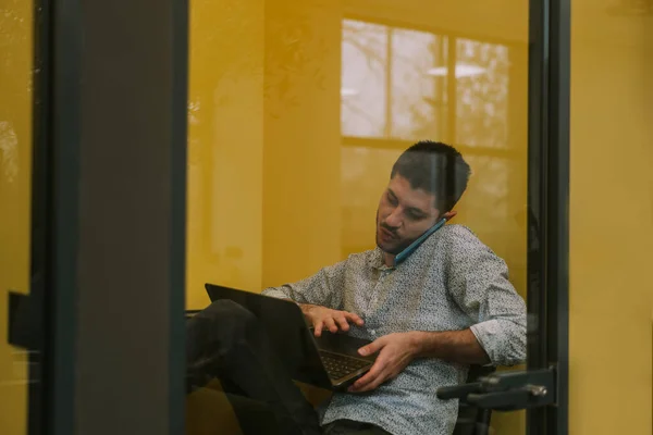 一位英俊的商人在办公室的隔音电话亭里与人私下交谈 一边在膝上型电脑上解决日常事务 — 图库照片