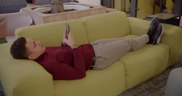 オフィスラウンジエリア内の居心地の良いソファで休んでいる若い男性労働者 — ストック動画
