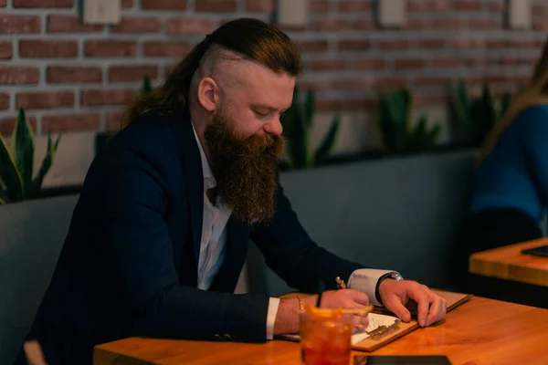 カフェでペンでノートに何か書いてる髭の男 — ストック写真