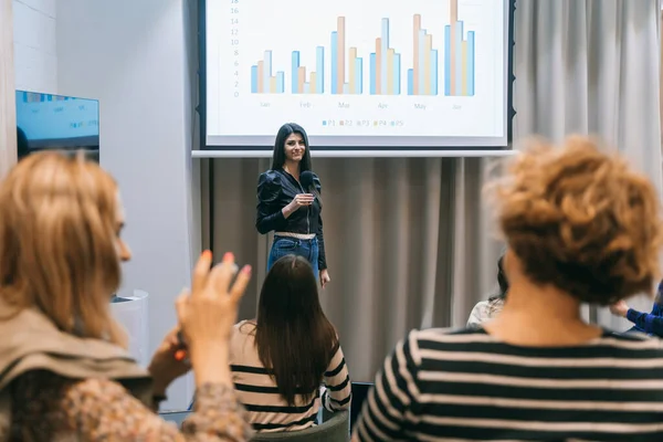 A shot of stunning female business coach giving speech to group of business employees. She is holding a microphone and looking at the camera
