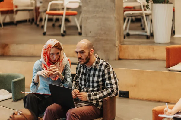 Woman in hijab working together on a new project with her hairless male partner. Young multicultural start-uppers working together at the office on a lap top