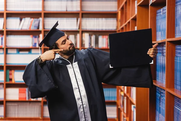 年轻而快乐的毕业生 戴着帽子 身穿长袍 指着他在图书馆的学士学位 — 图库照片