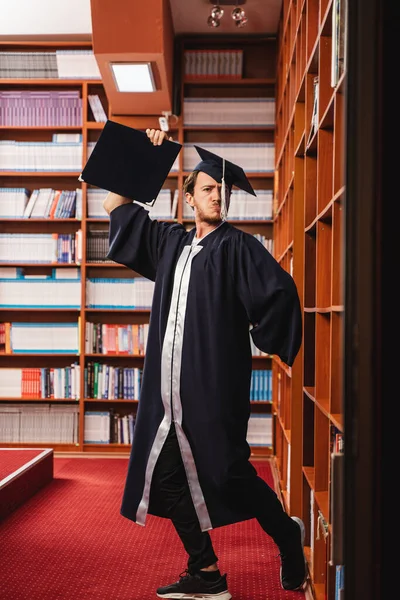 戴着帽子 身穿长袍的年轻而兴奋的毕业生在图书馆庆祝他获得学士学位 — 图库照片
