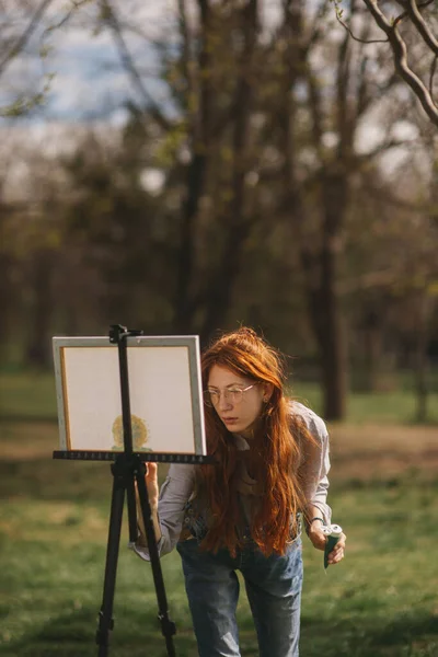 イーゼルの隣に立って公園で自然の中で絵を描いているジンジャーの女性 — ストック写真