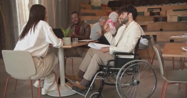 さまざまな起源と人種のビジネスの人々は 車椅子 オフィスラウンジエリア トラック移動で障害のある同僚のために包括的である間一緒に働いています — ストック動画