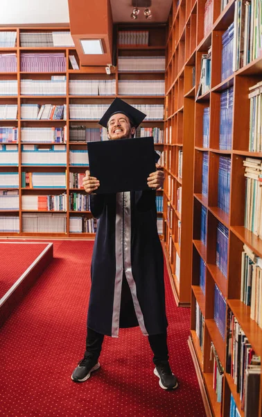 又兴奋又年轻的男子自豪地在图书馆里拿到了大学学位 — 图库照片