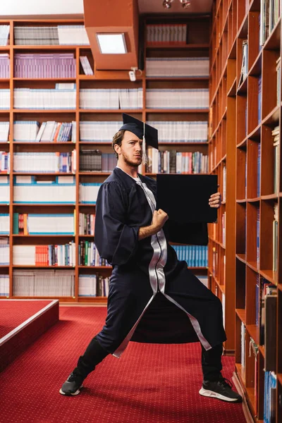 戴着帽子 身穿长袍的年轻而兴奋的毕业生在图书馆庆祝他获得学士学位 — 图库照片