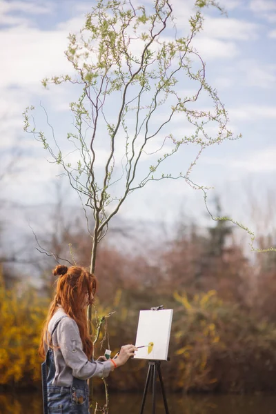 木の隣に立っている間 キャンバスに絵を描く美しいジンジャー女性 — ストック写真