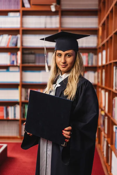 Young graduate holding her bachelor's degree and wearing a cap and gown inside a library during graduation