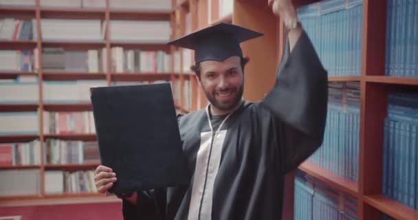 大学を卒業した後 誇らしげに図書館で卒業証書を保持したエキサイティングな学生の感覚 — ストック動画