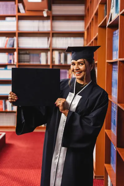 若くて幸せで美しい女性の卒業生は 図書館で彼女の大学の学位を保持しながらキャップとガウンを身に着けています — ストック写真