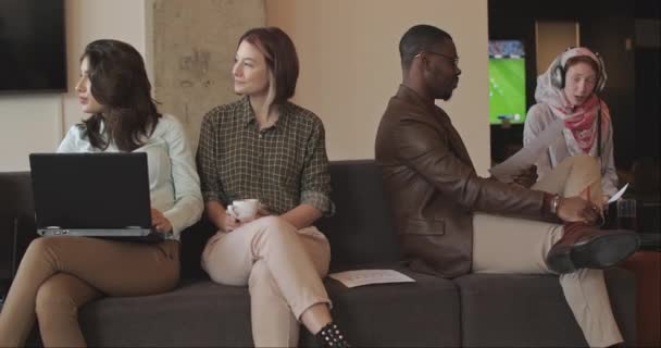 四个商人 一个黑人和一个女穆斯林 头戴头巾 一边谈论一个项目 一边笑着躺在舒适的办公室沙发上 其他同事在后面玩着模糊的游戏 相机卡车司机 — 图库视频影像