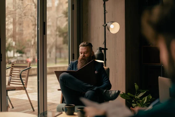 一个英俊的留着胡子的男人 双腿交叉地坐在咖啡店的椅子上 在记事本上写着什么 — 图库照片