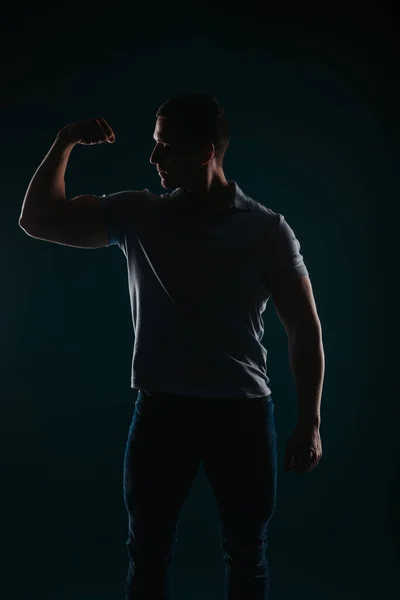Appealing and well-built man standing in a dark room and flexing his bicep